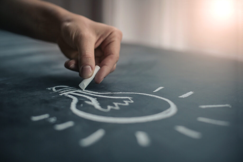 Kuvassa kädessä oleva liitu, jolla piirretään idealamppua tummalle taustalle.