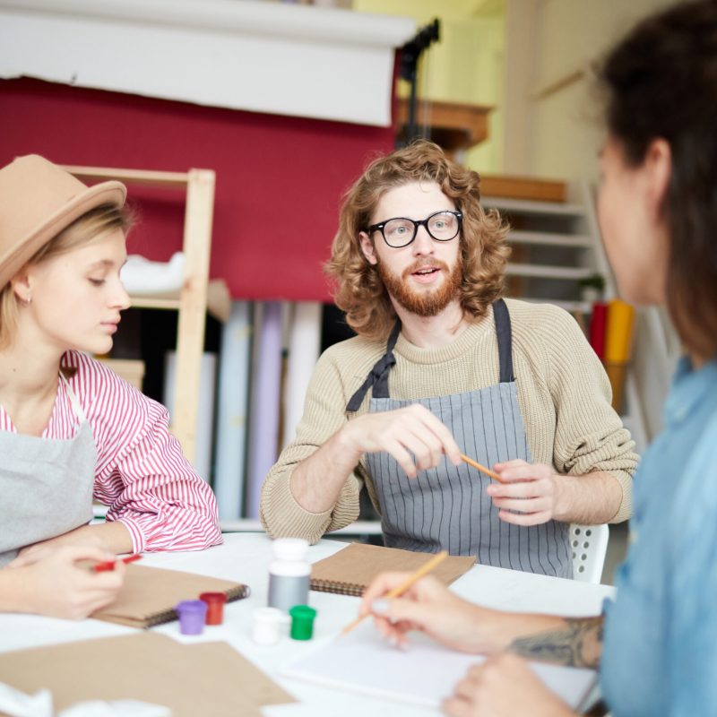 Young man and two girls sitting by desk and talking about principles of painting at working meeting